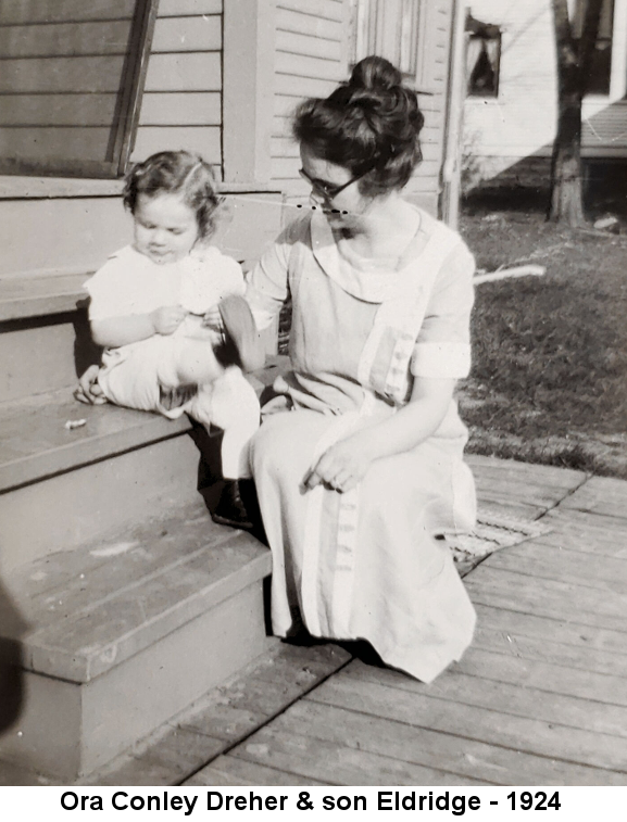 Black and white photo of a curly-haired toddler, Eldridge Conley Dreher sitting on the wooden steps of a frame house and kicking his right foot high; his mother Ora Conley Dreher, her hair piled high and wearing black-framed glasses, sits on the step next to him in a long light-colored dress.